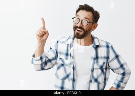 Good-looking creative and smart engineer in clenched shirt and glasses, looking and pointing up while smiling broadly and telling opinion to coworkers, discussing new invention over gray background Stock Photo
