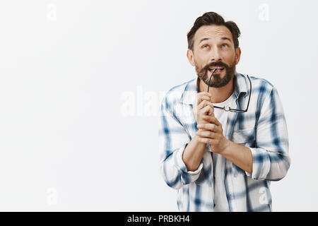 Charming strategist thinking up new plan. Portrait of dreamy friendly-looking male scientist in clenched shirt, biting rim of glasses while using imagination and gazing up to find idea in mind Stock Photo