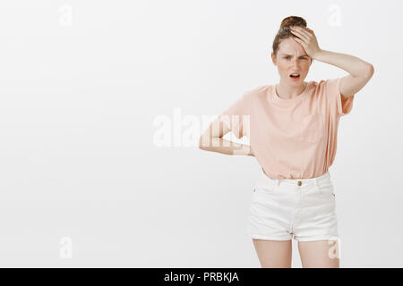 Young female feeling headache after partying all night, having hangover. Portrait of perplexed and confused woman with cute freckles, squinting and frowning, holding palm on forehead and dropping jaw Stock Photo