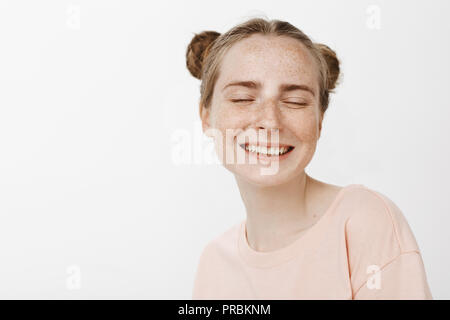 Close-up shot of carefree tender female with cute buns hairstyle and freckles, smiling joyfully with closed eyes and dreamy expression, fantasizing about prince on white horse over gray background Stock Photo