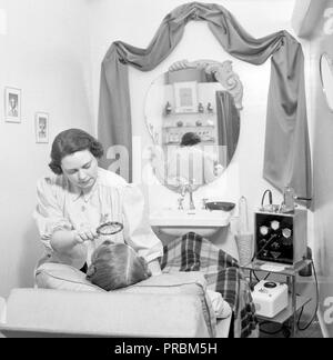 1950s beauty salon. A cosmetologist is examining a clients face with a magnifying glass. A state of the art electrical device with unknown function is standing on a side table.   Sweden 1950. ref 1519 Stock Photo