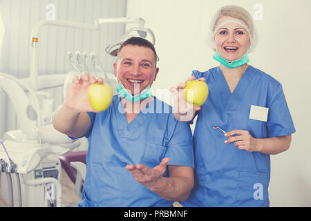 Two smiling professional dentists posing at dental clinic with fresh yellow apples in hands Stock Photo