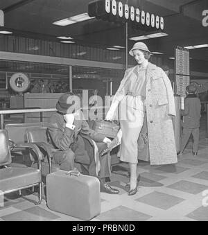 Couple in the 1950s. A young couple in an airport. She is wearing a fashionable two piece matching dress with an overcoat hanging over her shoulders. He is looking a bit bored waiting and is sitting resting his head on his hand and arm while reading a newspaper. Sweden 1956. Photo Kristoffersson Ref CB15-11 Stock Photo