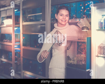happy european girl holding aquarian net and water container next to aquarium with colorful fish in aquarium shop Stock Photo