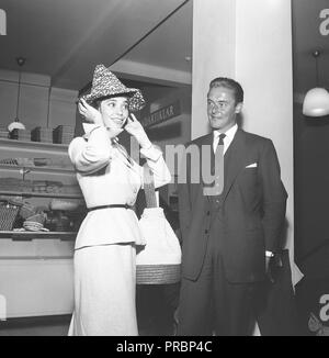 1950s fashion. Swedish actress Margit Carlqvist, born 1932 is trying on a hat while a man is standing beside her looking at her. Sweden 1956. Ref BX34-6 Stock Photo