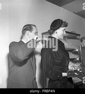 1950s fashion. A young fashion model is being prepared for the show, and a man is brushing off dust from the shoulders of the blouse she is wearing. Sweden 1951. Ref BE56-4 Stock Photo