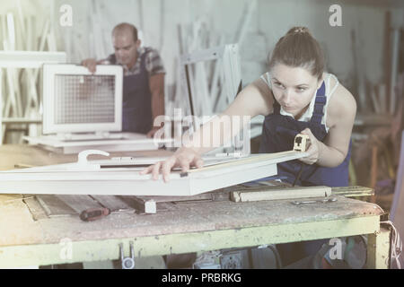 Woman 20-30 years old is checking dimensions of finished plastic window using measuring tape at factory. Stock Photo
