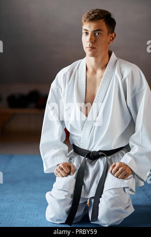 Karate man in kimono sits on knees on floor in martial arts gym Stock Photo