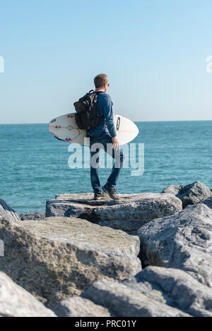 man standing on rocks holding a surfboard checking out the sea before surfing. Stock Photo