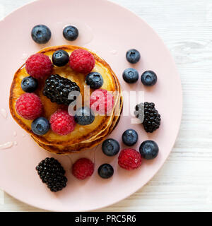 Pancakes with berries and honey on a pink plate on white wooden backfround, overhead view. Close-up. Stock Photo