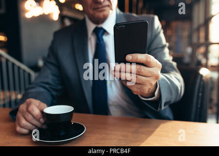 Close up of mobile phone in hand of a businessman with coffee on cafe table. Senior businessman using smartphone at coffee shop. Stock Photo