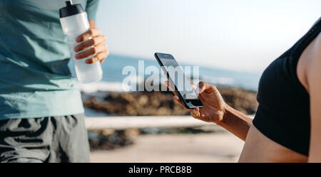 fitness couple standing outdoors and relaxing after workout. Couple taking a  break during workout checking mobile phone and drinking water. Stock Photo