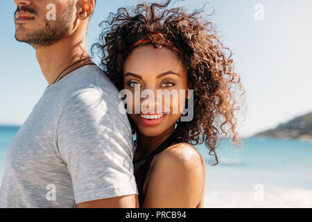 Side view of a smiling woman standing behind her boyfriend hugging him. Woman on vacation with her husband standing on beach. Stock Photo