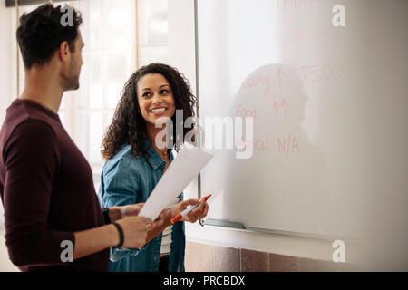 Office colleagues planning and discussing monthly budget on a whiteboard. Smiling businesswoman writing on the board while her colleague holds a paper Stock Photo