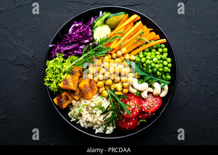 Bowl dish with brown rice, cucumber, tomato, green peas, red cabbage, chickpea, fresh lettuce salad and cashew nuts. Healthy balanced eating Stock Photo