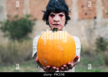 closeup of a scary disfigured man, wearing ragged and dirty clothes with blood stains, holding a pumpkin in his hands, in front of an abandoned house Stock Photo