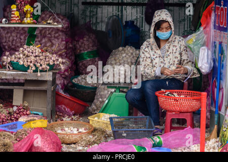 A woman wearing a dust mask peels thousands of bulbs and cloves of garlic on the street in Hanoi, Vietnam. Stock Photo