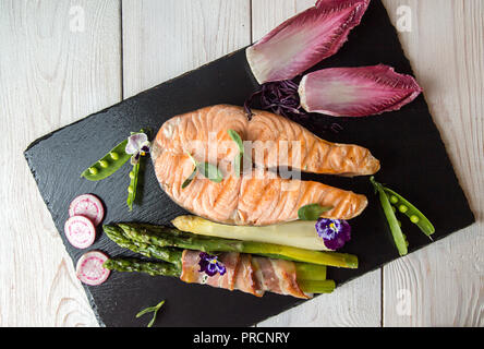 Baked in oven grilled salmon, stick of green asparagus wrapped in bacon on white background. Overhead. Copy space. Stock Photo