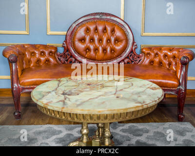 Vintage luxury red brown leather sofa with marble and brass round table on gray carpet and wooden floor on light blue and golden edge wall background. Stock Photo