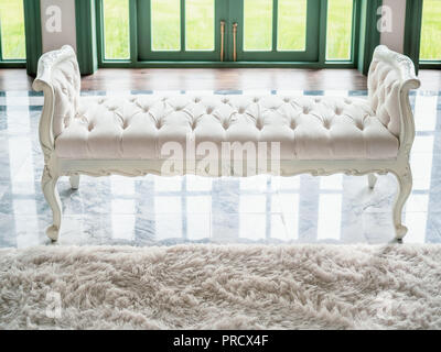 Classic vintage white tufted bench or bedroom bench on marble floor with white soft fur carpet in contemporary house near green door. Stock Photo
