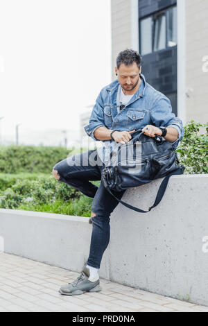 Young man rummaging in black sport bag and sitting on cityscape background. Stock Photo
