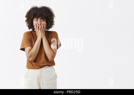 Portrait of panicking timid and insecute cute dark-skinned female model with curly hair holding palms on mouth not to scream feeling anxious or nervous staring right over gray background Stock Photo