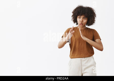 Kiai do not mess with me I know martial arts. Portrait of funny and cute playful African American curly-haired woman in trendy t-shirt raising palms in kung fu pose folding lips over gray wall Stock Photo