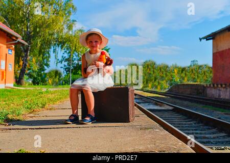 Cute girl holding stuffed animal on a railway station, waiting for the train with vintage suitcase. Traveling, holiday and chilhood concept. Travel in Stock Photo