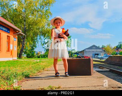 Cute girl holding stuffed animal on a railway station, waiting for the train with vintage suitcase. Traveling, holiday and chilhood concept. Travel in Stock Photo