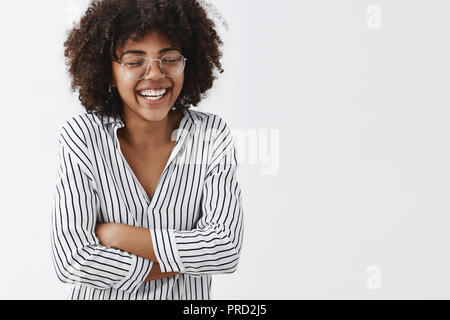 Belly aching from laughter. Amused and carefree attractive african american woman in striped blouse and glasses closing eyes laughing out loud and holding hands on chest closing eyes having fun Stock Photo