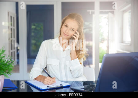 Portrait of beautiful young assistant businesswoman making call while sitting at desk and making notes. Stock Photo