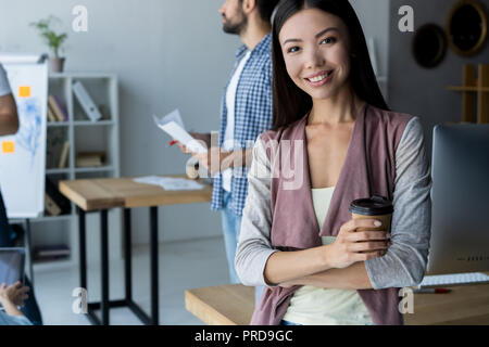 Confident and successful leader. Confident young woman keeping arms crossed and looking at camera with smile while her colleagues working in the background. Stock Photo