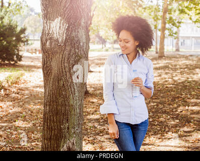 Portrait of smiling young black woman holds bottle of water in nature, afro hair style ,wear blue shirt, sunlight flare in background
