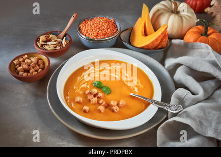 Pumpkin and red lentil creme soup in ceramic bowl seasoned with basil, cream and croutons, with ingredients and seasoning around Stock Photo