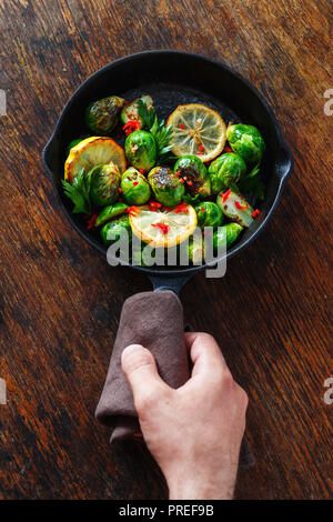 Male hand holding frying pan with fried Brussels sprouts on dark wooden background top view Stock Photo