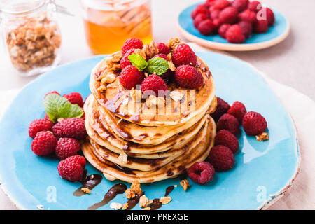 Stacked american pancakes with raspberry and chocolate syrup, sprinkled with granola and decorated with mint leaves. Served on blue plate, on light st Stock Photo