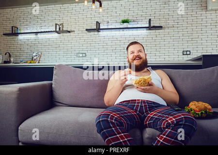 Fat funny man in pajamas eating a burger at home. Stock Photo