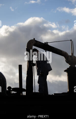Close up of railway steam train crew, in sky silhouette, standing on vintage UK locomotive at water stop, engine taking on water, early evening. Stock Photo