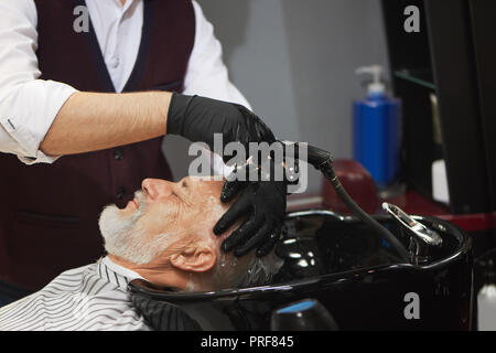 Side view of old man with grey hair holding head in black wash basin. Unrecognizable barber in black gloves washing head of his client. Concept of barbershop procedures. Stock Photo