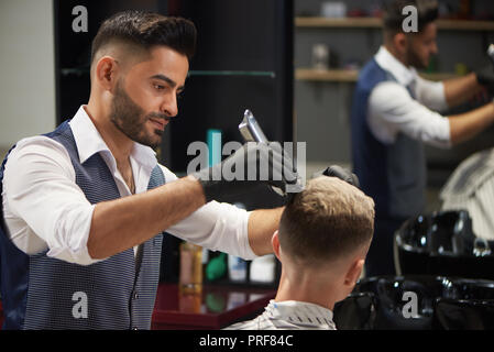 Concentrated male hairdresser holding trimmer in hands and trimming haircut of client. Bearded master wearing white shirt, vest and black gloves, while working in barbershop. Stock Photo