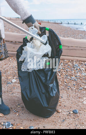 A woman using a litter picker puts plastic waste found on the beach into a black rubbish bag during a community beach clean up. Stock Photo