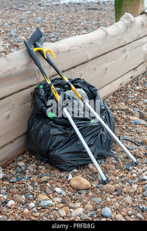 Beach clean up. A black bag full of rubbish collected from the beach and two litter pick up tools leaning on a wooden groyne on the beach. Stock Photo