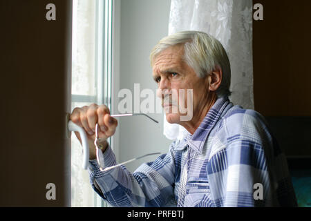 portrait of a gray-haired man 60 years old. A man looks out the window. The man has poor eyesight, he wears glasses Stock Photo