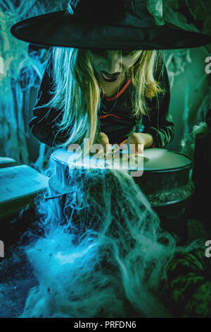 Young angry witch with awfully face tells evil words, and putting bones in cauldron with poison potion. Stock Photo