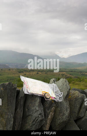 Ordnance Survey map in a plastic case with a compass on a drystone wall in the Lake District, overlooking a cloudy and misty Broughton Moor in Furness. Stock Photo