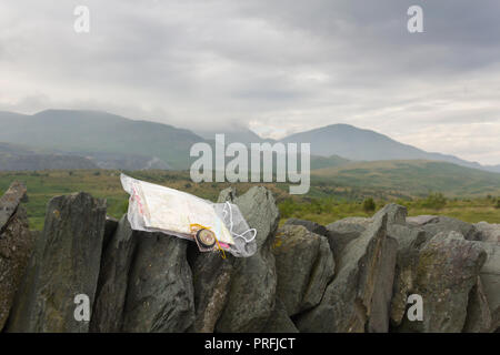 Ordnance Survey map in a plastic case with a compass on a drystone wall in the Lake District, overlooking a cloudy and misty Broughton Moor in Furness. Stock Photo