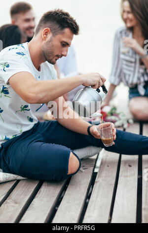 Young man pours tea into cup on picnic sitting on wooden pier. Camping food concept Stock Photo