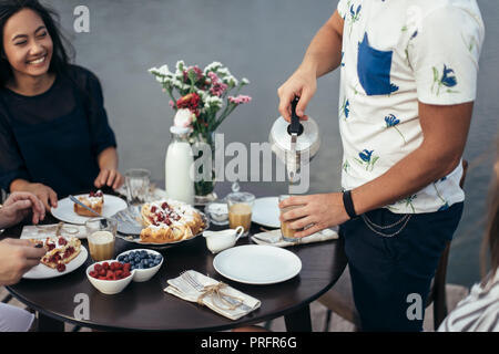 Young man with teapot pouring tea into cup during friendly breakfast by river. guy in white shirt filling glass with masala. Culinary delights in fres Stock Photo