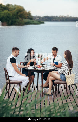Group of friends enjoying meal on picnic in river beach pier. Happy people having fun on nature Stock Photo
