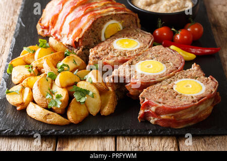 Delicious bacon wrapped meat loaf with egg served with potato wedges and sauce close-up on a slate plate on the table. horizontal Stock Photo